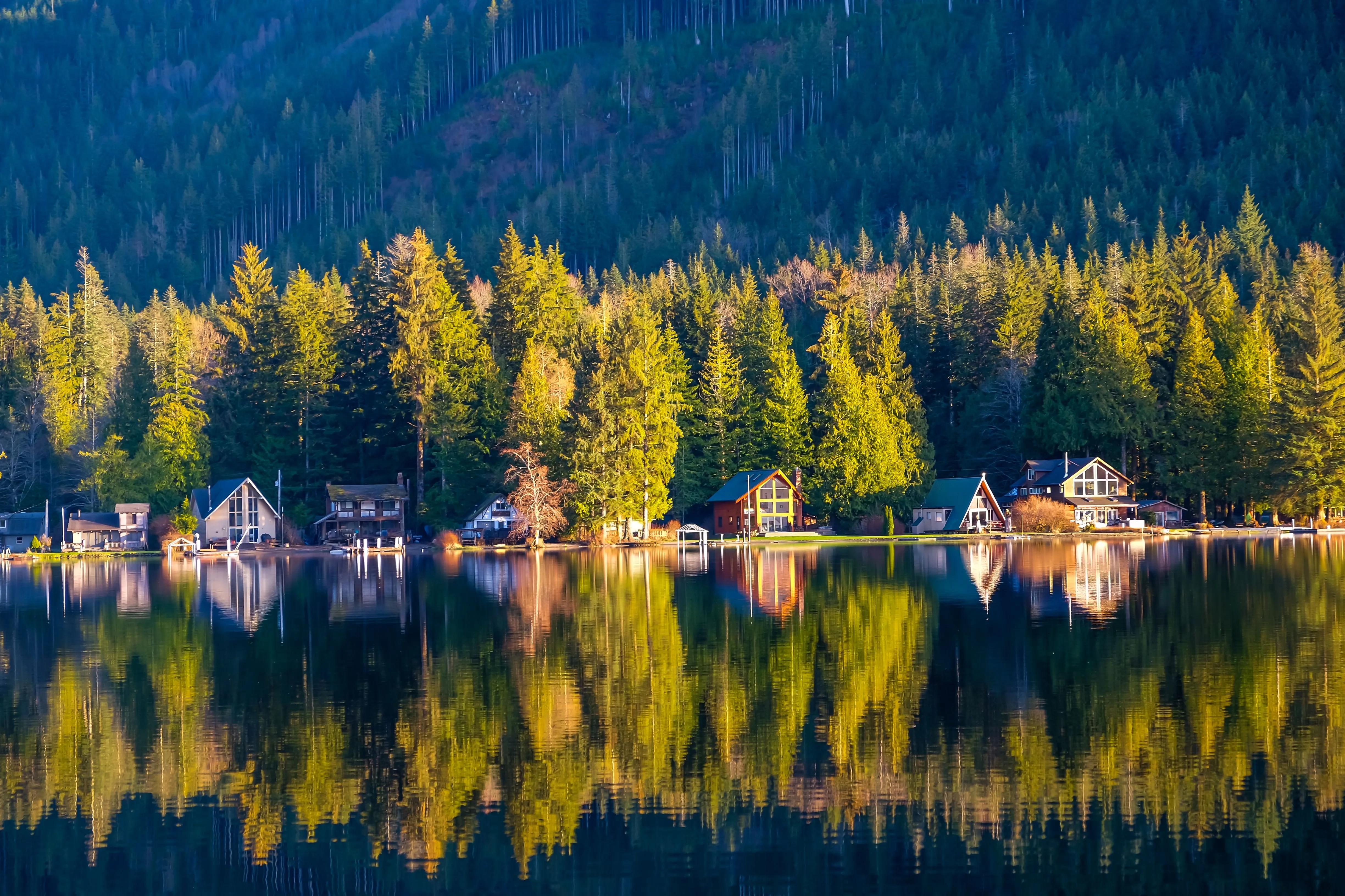 Homes along a still lake shore, mirrored in the water under autumn timber