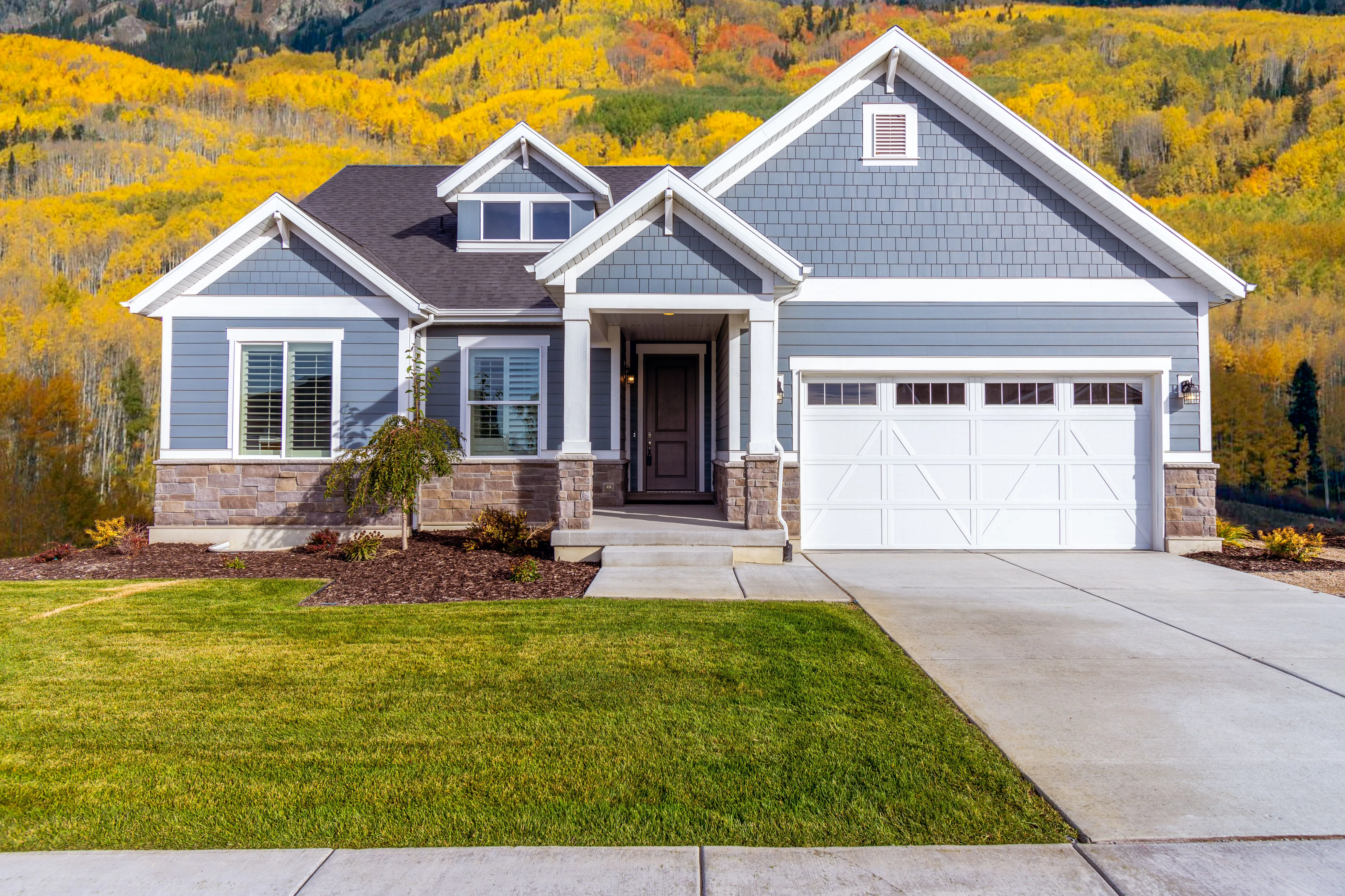 A newly finished blue-grey home with a white-trimmed porch and double garage, hillsides of golden aspens behind it