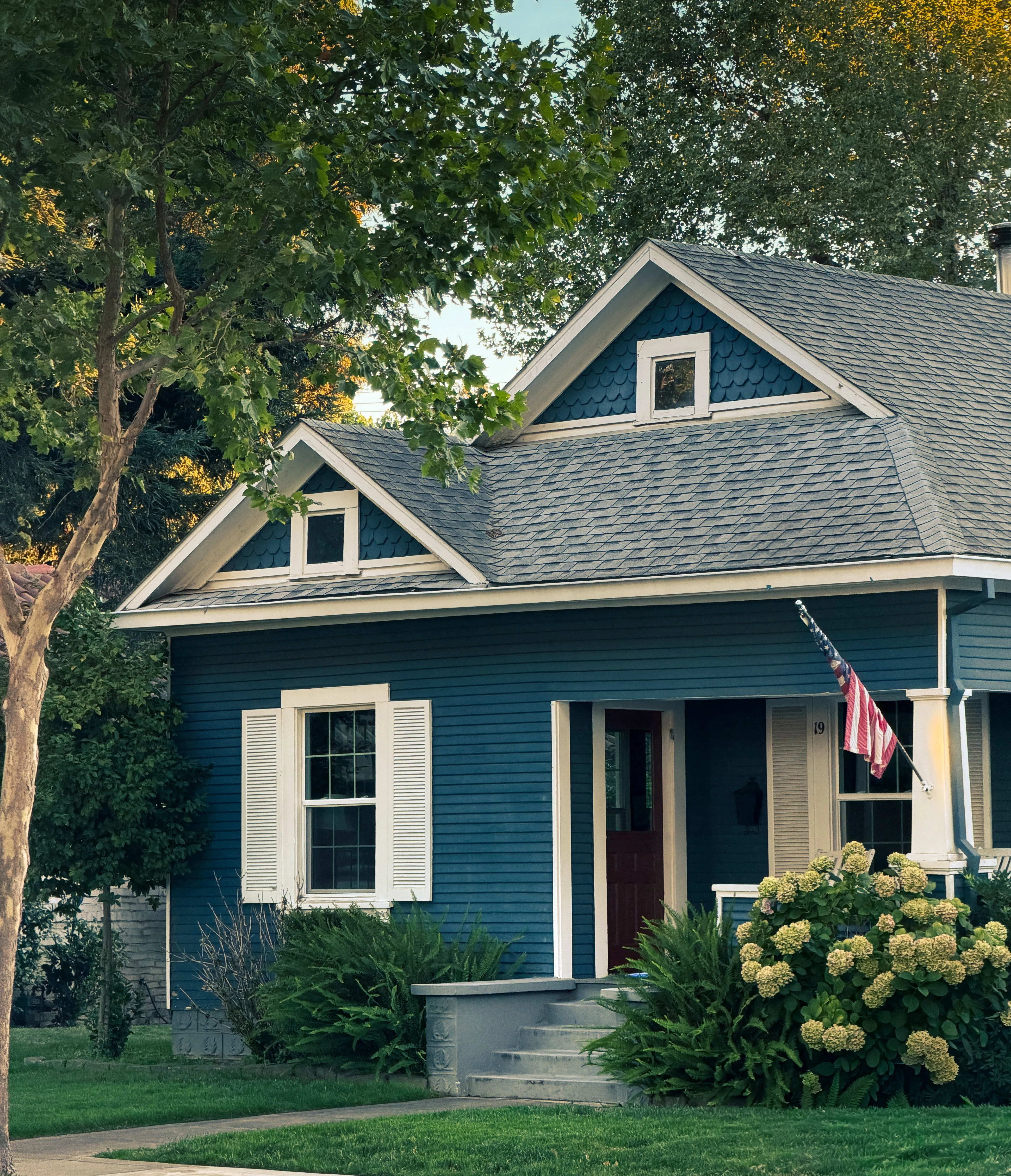 A blue cottage-style house with white trim and a covered front porch, hydrangeas by the steps