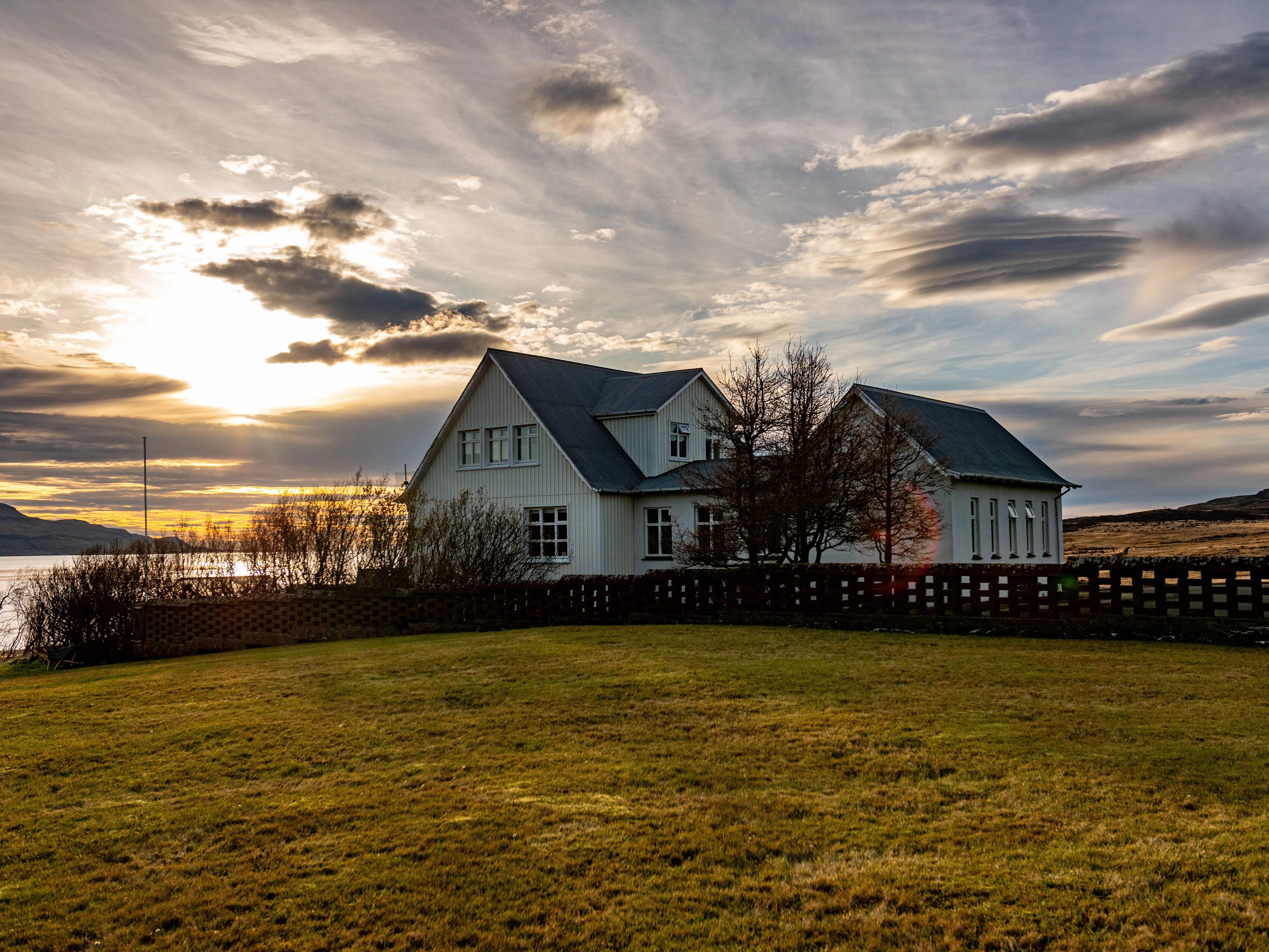A white farmhouse-style home across an open field at sunset, a wide sky of gold-edged clouds above it