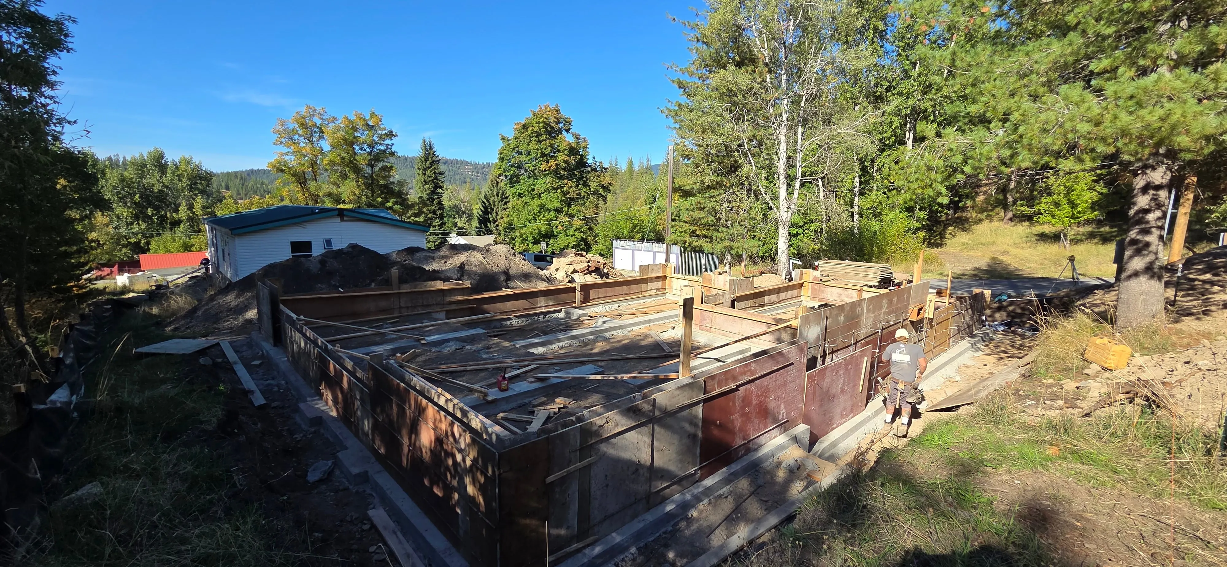 The poured foundation walls of one of Jared's builds, a neighbouring house and pines beyond