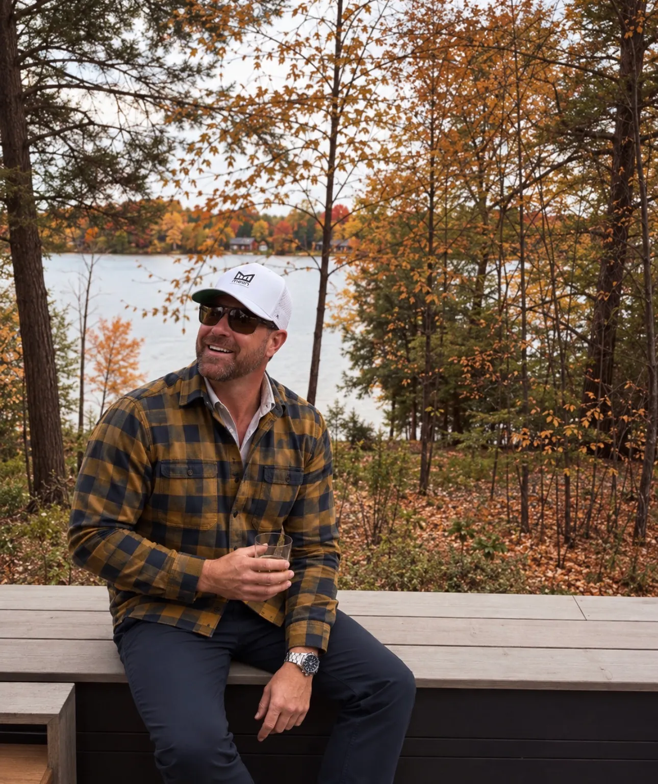 Jared Ban sitting on a deck rail in a plaid flannel shirt, autumn woods behind him