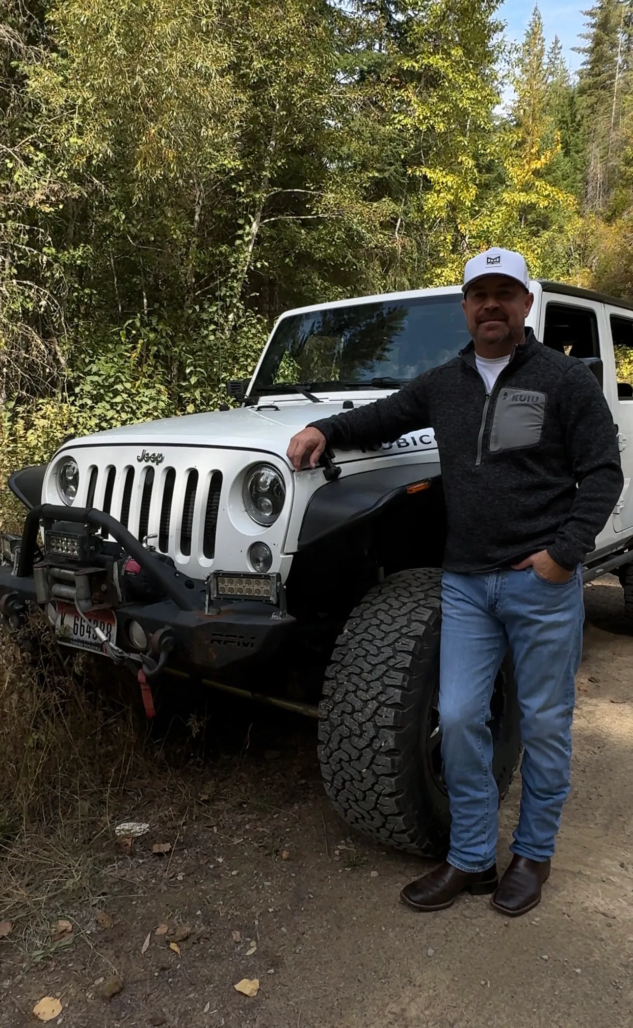 Jared Ban leaning against the hood of his white Jeep on a wooded trail