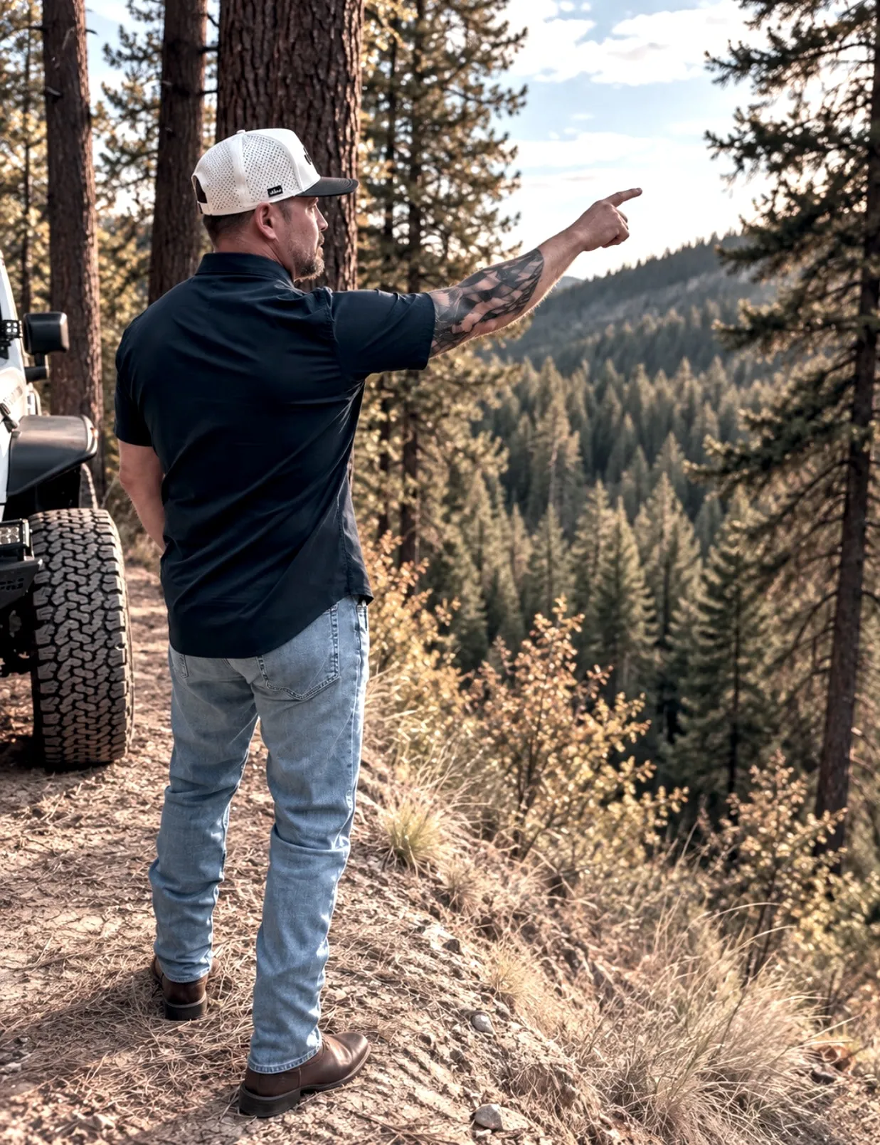 Jared Ban pointing out across a valley from beside his Jeep, timbered hills beyond