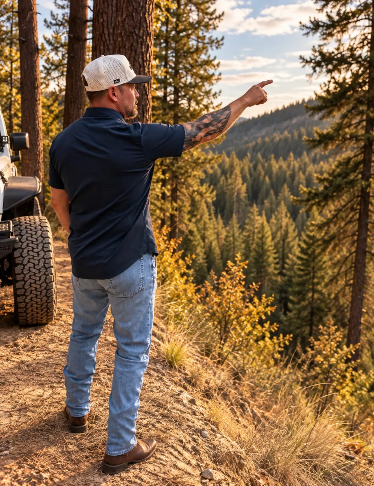 Jared Ban pointing out across a valley from beside his Jeep, timbered hills beyond