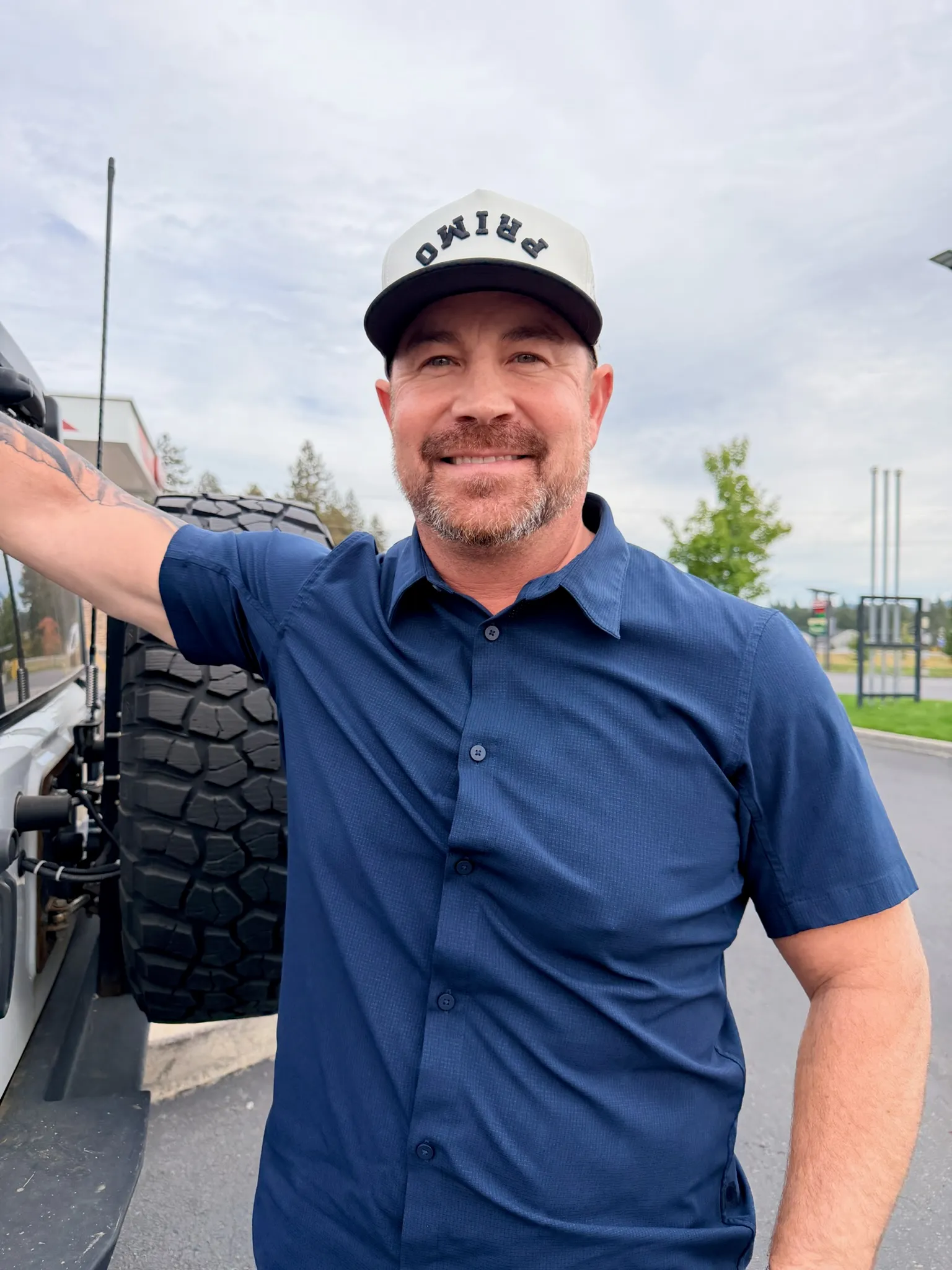 Jared Ban smiling, one arm resting on the spare wheel of his Jeep, on a bright overcast day