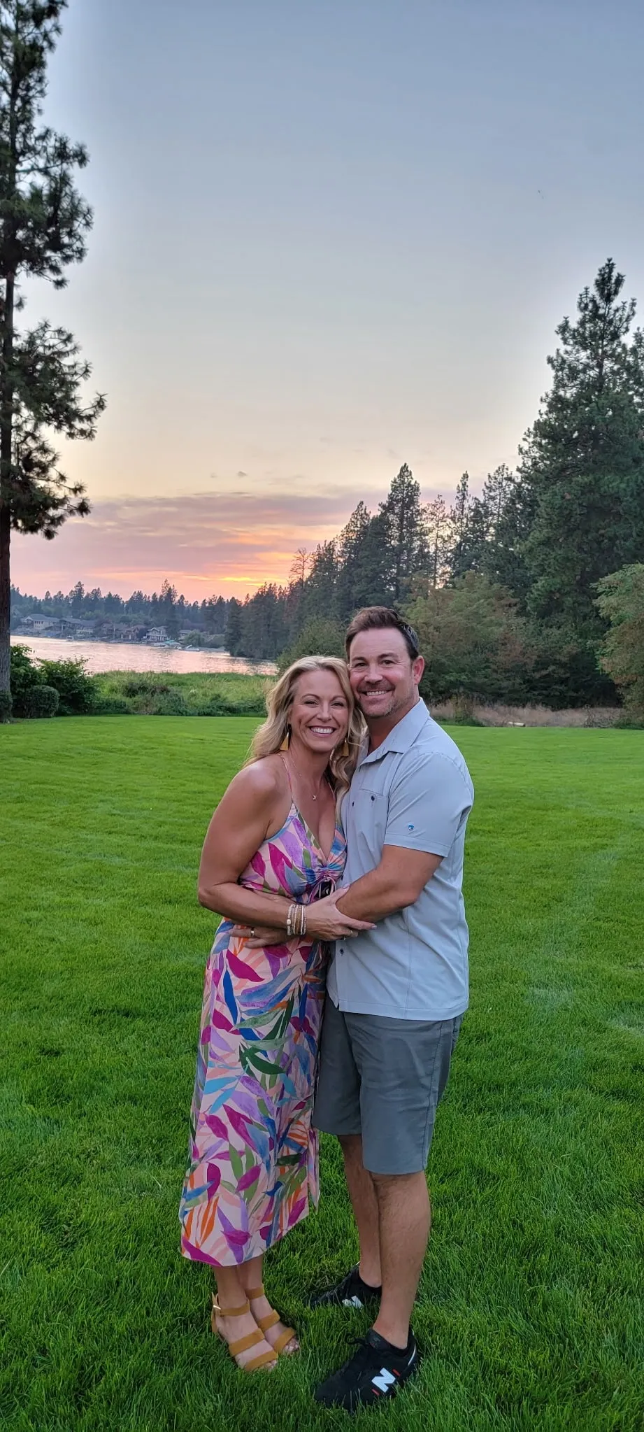 Jared Ban and his wife Shadra arm in arm on a lawn at sunset, a lake and pines behind them