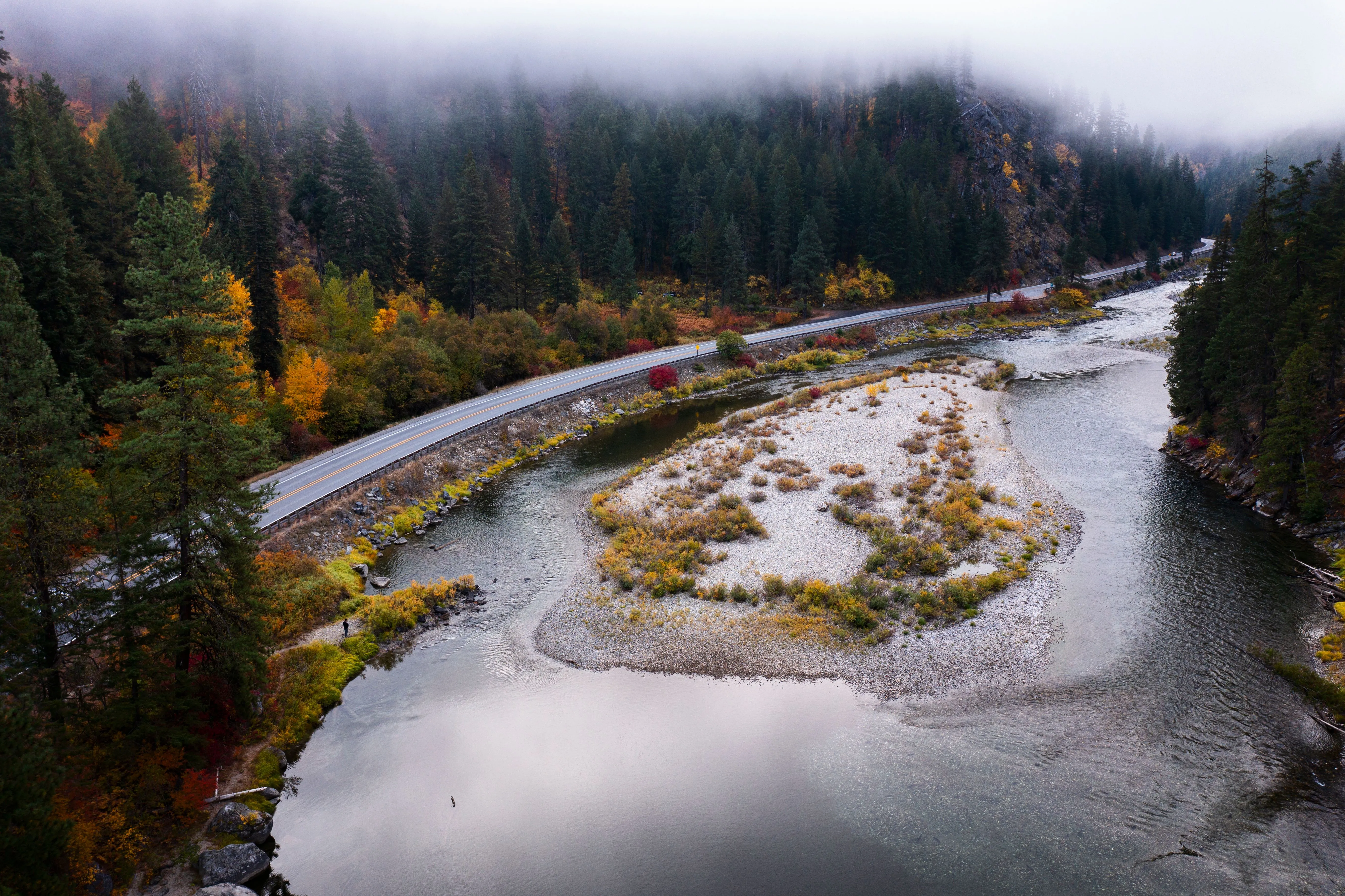 A river bends around a gravel bar past fog-covered forested hills and a mountain highway