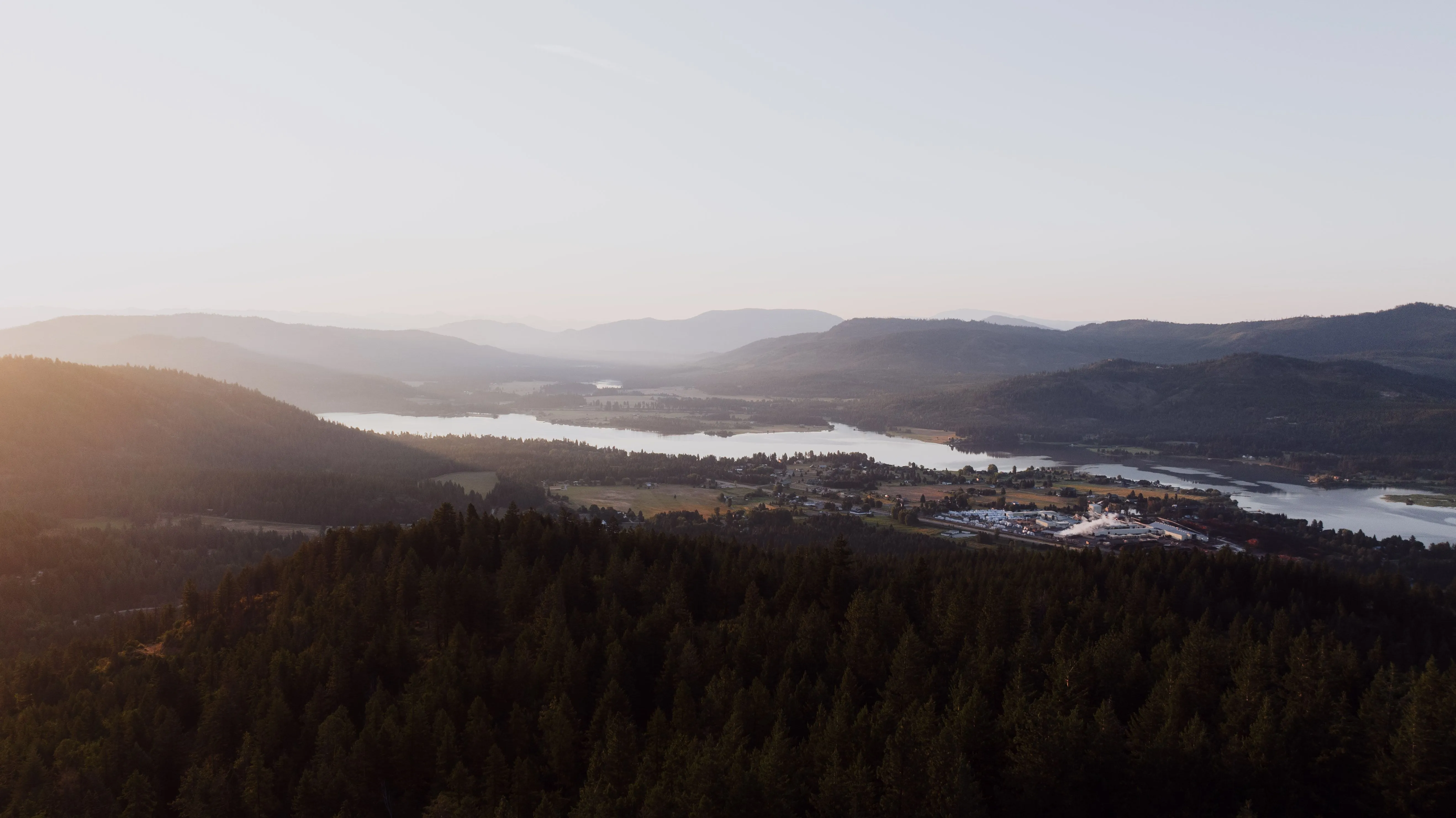 An aerial view of forested ridgelines falling away to a long lake valley in hazy golden light
